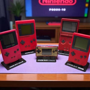 May include: A collection of red Game Boy consoles, including a Game Boy Color, Game Boy, Game Boy Advance SP, and Game Boy Pocket, along with a gold Game Boy Micro. Each console is displayed on a black stand with its name.