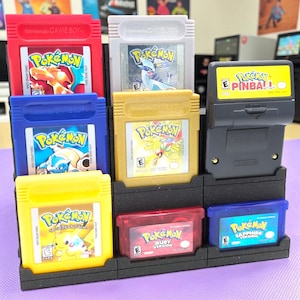 May include: A black display stand holding various Game Boy Pokémon game cartridges. The cartridges are in red, blue, yellow, gold, and gray. The games include "Pokémon," "Pokémon Pinball," and "Pokémon Ruby Version."