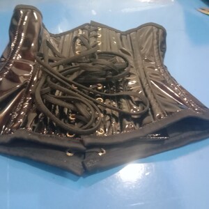 May include: A black patent leather corset with black lacing and gold grommets. The corset is unlaced and laid flat on a blue surface.