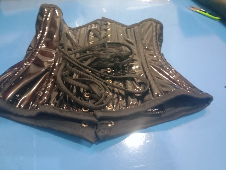 May include: A black patent leather corset with black lacing and gold grommets. The corset is unlaced and laid flat on a blue surface.