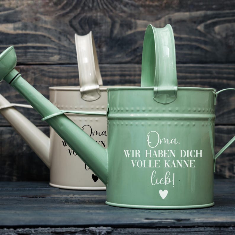 May include: Two metal watering cans, one sage green and one off-white, with arched handles and long spouts. The green can has white text that reads "Oma. Wir haben dich volle Kanne lieb!" with a heart. The off-white can has similar text.
