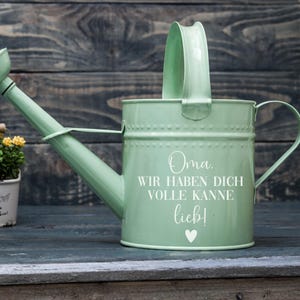 May include: A mint green metal watering can with a curved spout and a large handle. The can has the words "Oma, WIR HABEN DICH VOLLE KANNE lieb!" and a heart symbol in white. The watering can is on a wooden surface.