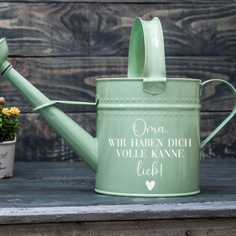 May include: A mint green metal watering can with a curved spout and a large handle. The can has the words "Oma, WIR HABEN DICH VOLLE KANNE lieb!" and a heart symbol in white. The watering can is on a wooden surface.