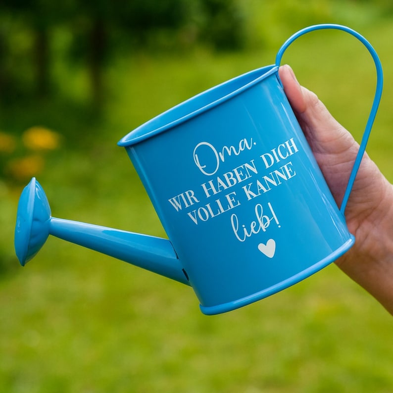 May include: A light blue metal watering can with a long spout and a curved handle. The can has white text that reads "Oma. Wir haben dich volle Kanne lieb!" with a heart symbol. The background is blurred green.