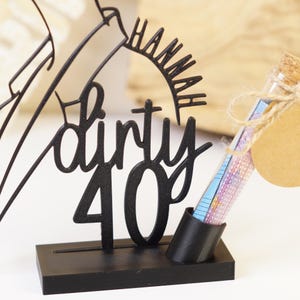 以下が含まれることがあります： 「dirty 40」の文字と「Hannah」の名前が入った黒い金属製のバースデーケーキトッパー。トッパーの横には、コルク栓とギフトタグが付いた小さなガラス管があります。チューブにはカラフルな紙が入っています。トッパーは黒いベースの上にあります。