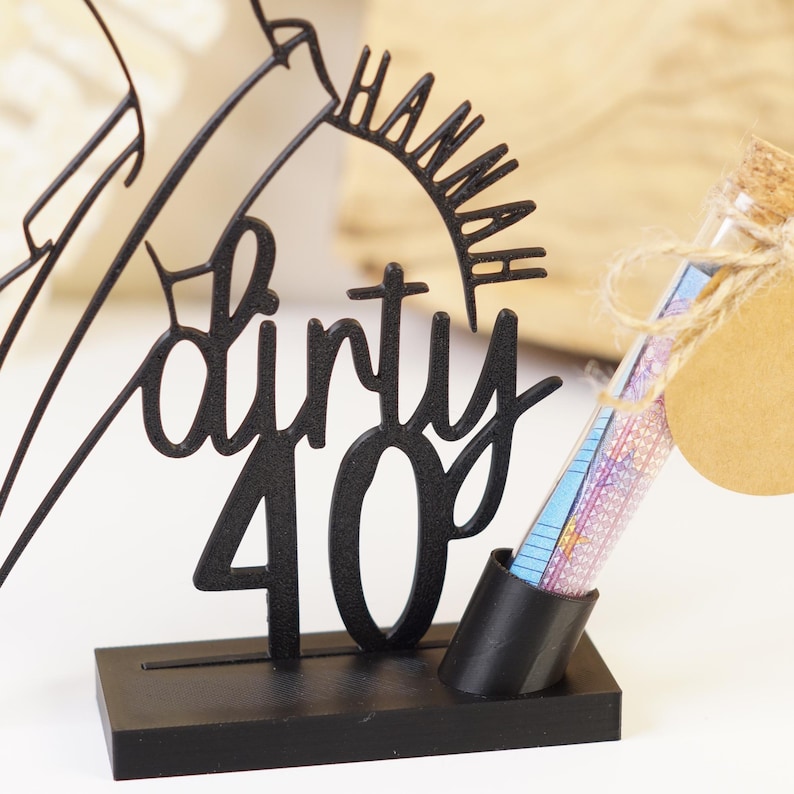 以下が含まれることがあります： 「dirty 40」の文字と「Hannah」の名前が入った黒い金属製のバースデーケーキトッパー。トッパーの横には、コルク栓とギフトタグが付いた小さなガラス管があります。チューブにはカラフルな紙が入っています。トッパーは黒いベースの上にあります。