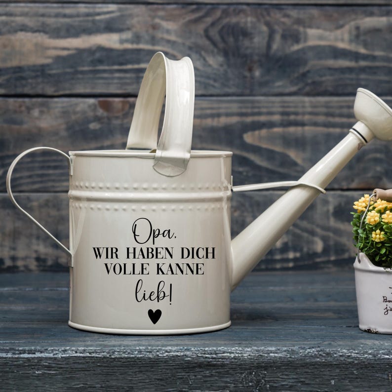 May include: Cream-colored metal watering can with a curved handle and spout. The can has the text "Opa. Wir haben dich volle Kanne lieb!" and a heart symbol. The watering can is sitting on a dark blue wooden surface.