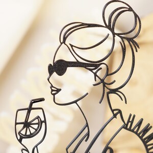 以下が含まれることがあります： サングラスをかけた女性がストローで飲み物を飲んでいる黒い金属製のシルエット。女性の髪はまとめられています。飲み物には柑橘系の果物のスライスが見えます。「HAMM」という言葉が部分的に見えます。