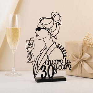 Könnte beinhalten: Ein schwarzer, lasergeschnittener Acryl-Tortentopper mit der stilisierten Umrisszeichnung einer Person mit Dutt und Sonnenbrille, die ein Cocktailglas hält. Der Text lautet "HANNAH" und "cheers to 30 years" in Block- und Schreibschrift.