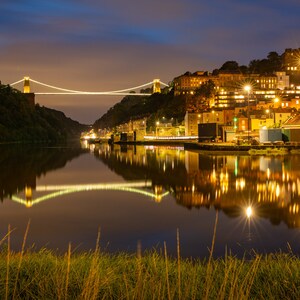 Puede incluir: Un puente colgante iluminado por la noche con su reflejo en el agua. El puente está iluminado con luces blancas y los edificios al otro lado del río están iluminados con luces amarillas.