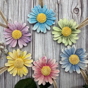 May include: Six daisy flower pendants in various colours, including blue, purple, yellow, pink, and green, each with a yellow centre. The flowers are attached to beige ribbons, displayed on a wooden background.