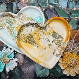 May include: Two heart-shaped trinket dishes with gold leaf detailing. One is pale blue, the other is cream and orange. Two decorative flower pendants with twine are also present. The items are on a mosaic tile surface.