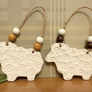 May include: Two white ceramic sheep ornaments with a spiral pattern, each with a jute string for hanging. The ornaments have wooden and white beads. The sheep are displayed on a wooden surface with green leaves.
