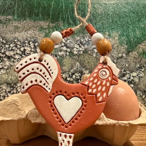 May include: A terracotta-coloured rooster-shaped ornament with white accents and a heart design. The ornament is strung with wooden beads and a jute cord. An egg is in an egg carton in the background.