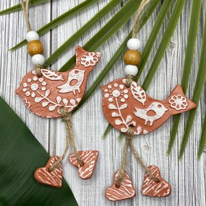 May include: Two terracotta bird ornaments with white floral designs and a small bird on each. Each bird has a string with three small terracotta hearts hanging from it.