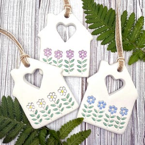 May include: Three white ceramic house ornaments with heart-shaped cutouts and floral designs. Each ornament has a different flower colour: purple, white, and blue. They hang from twine cords, and are displayed on a wooden surface.