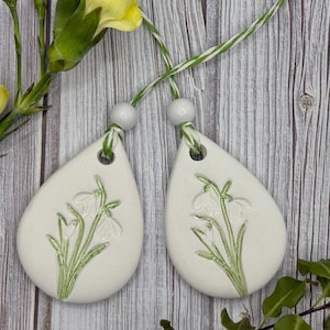 May include: Two off-white ceramic teardrop ornaments, each featuring a raised snowdrop flower design in green. They hang from green and white cords with white beads. The background is a wooden surface with yellow flowers.