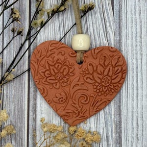 May include: A terracotta heart-shaped ornament with an embossed floral pattern, suspended by a beige cord and a white bead. The ornament is set against a weathered, light-toned wooden backdrop, with dried flowers.