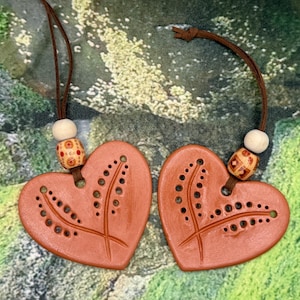 May include: Two terracotta-coloured heart-shaped ornaments with brown leather cords. Each heart features a cut-out design and is adorned with a wooden bead and a decorative bead. The background is a blurred green and brown.