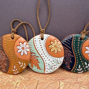 May include: Three ceramic pendant necklaces with leather cords. Each pendant is round with a floral and leaf design. The colour palette includes orange, brown, white, and green. The pendants are handmade and suitable for everyday wear.