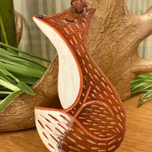 May include: A hand-carved wooden fox ornament with a leather cord for hanging. The fox is painted in shades of brown, with a white chest and belly. The fox has a black nose and closed eyes.