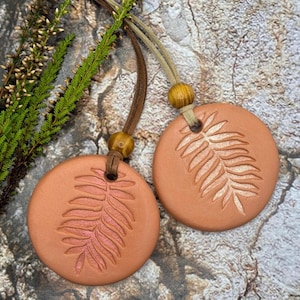 May include: Two terracotta-coloured round pendants with a fern leaf design, suspended by leather cords and wooden beads. The pendants are displayed on a textured, neutral-toned surface, with sprigs of greenery.