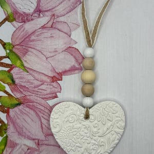 May include: White heart-shaped ornament with a floral embossed design, suspended by a jute and wooden bead strand. The background features a watercolor floral design in shades of pink and green.
