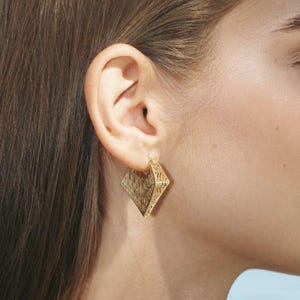 May include: A person's profile featuring a gold diamond-shaped earring. The earring has a textured design and is suspended from the ear. The person has long brown hair and is wearing a patterned top. The background shows a blurred ocean view.