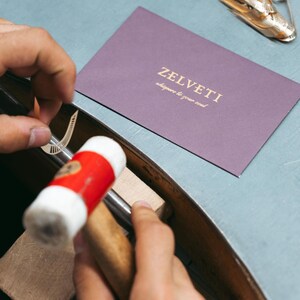 May include: A goldsmith at work, shaping a small metal component with a hammer. The hammer has a red and white handle. A purple card with gold text reading "ZELVETI whispers to your soul" is visible in the background.