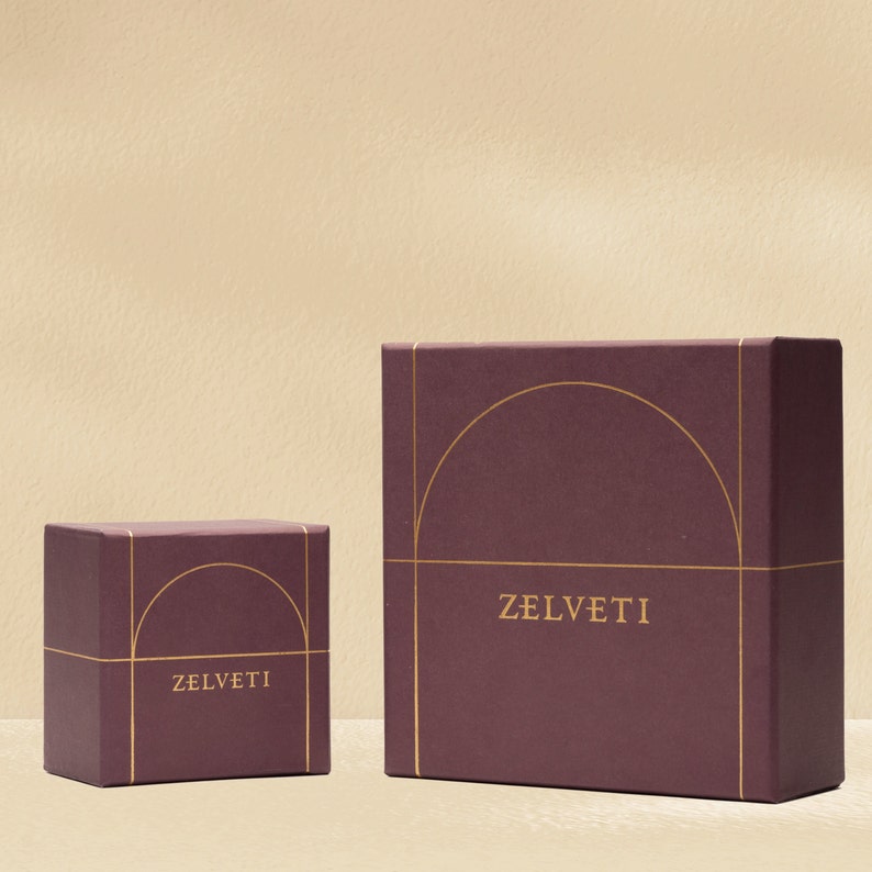 May include: Two maroon gift boxes with gold detailing. The boxes are rectangular, with the word "ZELVETI" printed in gold on the front. The larger box features a gold arched design. The boxes are on a cream-coloured surface.