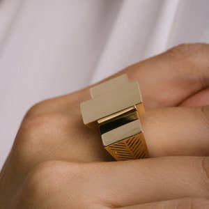 May include: A gold ring with a geometric design. The ring has a square shape with a smaller square on top. The smaller square has a textured surface.