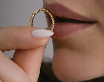 May include: A gold-colored twisted band ring. The ring is being held by a finger with a white nail polish.