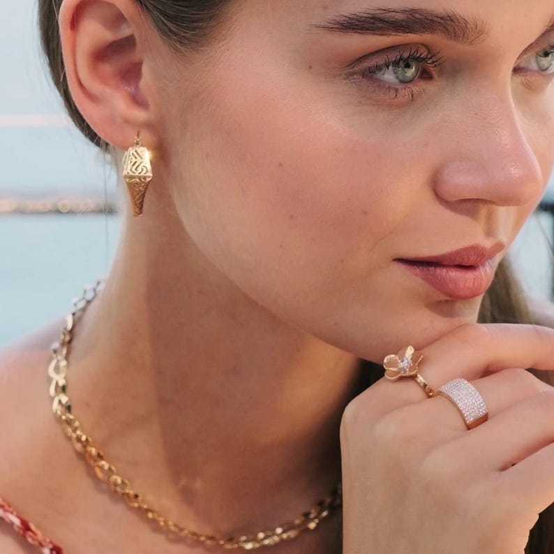 May include: A woman wearing gold jewellery, including an ice cream cone-shaped earring, a necklace, and two rings. One ring is a flower design, and the other is a wide band with small stones. The jewellery is set against a soft background.