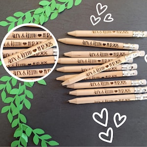 May include: Wooden pencils engraved with "ALEX & KEITH 11.12.2025" and a heart symbol. The pencils are arranged on a dark surface, with decorative green leaves and white heart illustrations.