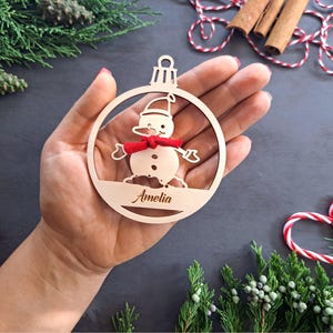 May include: A wooden Christmas ornament shaped like a snowman with a red scarf. The ornament is personalized with the name "Amelia".