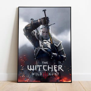 May include: Framed poster featuring a detailed illustration of a character from "The Witcher 3: Wild Hunt" video game. The character is holding a sword, with the game's title prominently displayed in white and red text. The background is a dark, smoky scene.