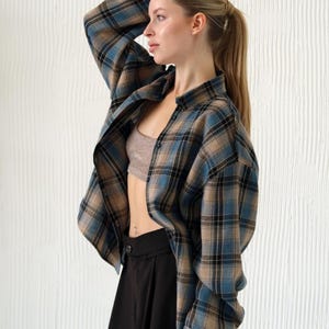 May include: A blue, tan, and black plaid flannel shirt is worn open over a neutral-colored sports bra. The shirt is paired with black high-waisted trousers. The model has blonde hair pulled back in a ponytail.