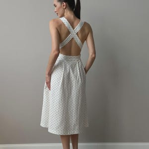 Puede incluir: Un vestido midi blanco con estampado de lunares. El vestido presenta un dise&ntilde;o de espalda cruzada y una falda acampanada. La modelo lleva zapatos negros. El vestido es una opci&oacute;n elegante para un d&iacute;a de verano.