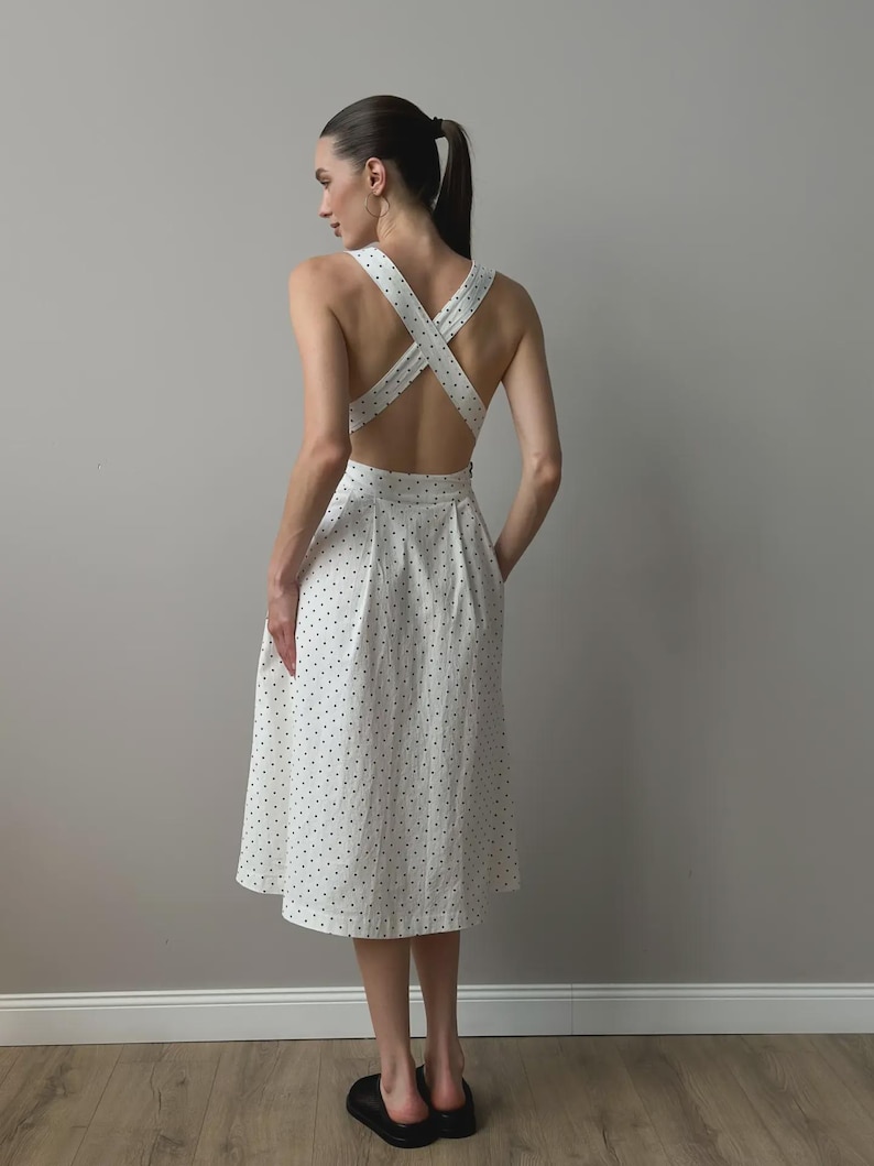 Puede incluir: Un vestido midi blanco con estampado de lunares. El vestido presenta un dise&ntilde;o de espalda cruzada y una falda acampanada. La modelo lleva zapatos negros. El vestido es una opci&oacute;n elegante para un d&iacute;a de verano.