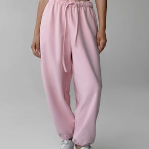 May include: Light pink sweatpants with a drawstring waist and elastic cuffs. The pants are made of a soft material and have a relaxed fit. The model is wearing white sneakers. The overall theme is casual wear.