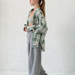 May include: A casual outfit featuring a green and white plaid shirt, a gray sports bra, and high-waisted gray trousers. The ensemble is completed with brown and black shoes. The background is a textured white wall.