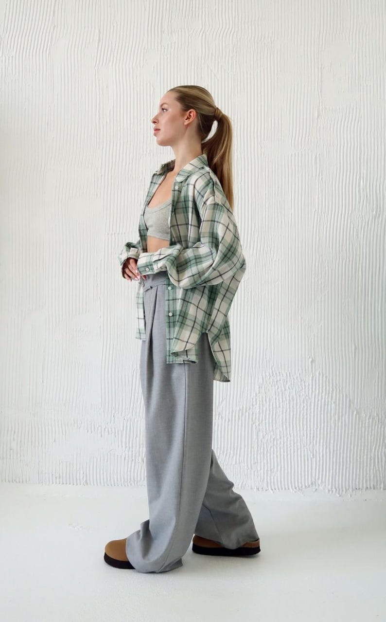 May include: A casual outfit featuring a green and white plaid shirt, a gray sports bra, and high-waisted gray trousers. The ensemble is completed with brown and black shoes. The background is a textured white wall.