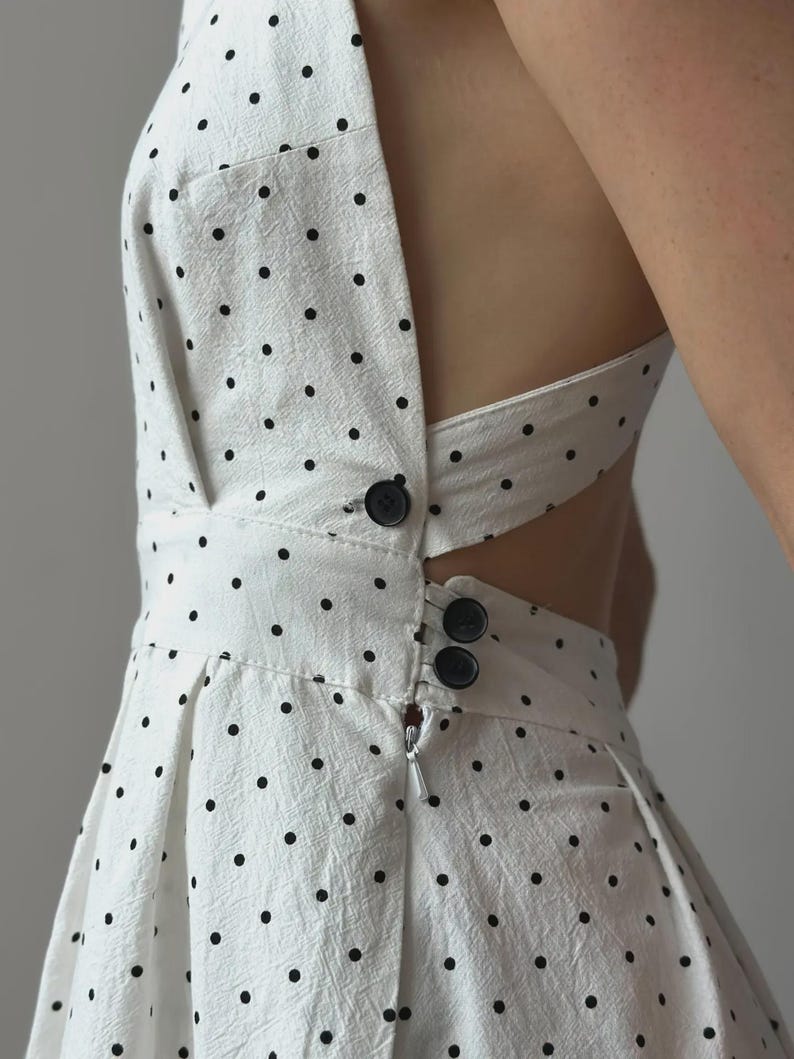 Puede incluir: Vestido blanco con lunares negros. El vestido presenta un cuello halter, un corpi&ntilde;o ajustado y una falda acampanada. Tiene cierre de cremallera y botones negros. El vestido est&aacute; hecho de una tela texturizada.