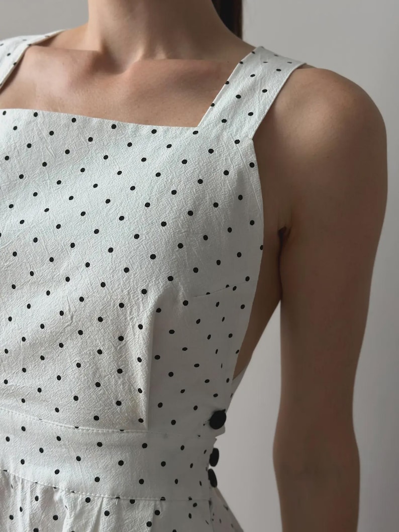 Puede incluir: Vestido blanco con lunares negros. El vestido presenta un escote cuadrado, tirantes anchos y un corpi&ntilde;o ajustado. Tiene un cierre de botones en el lateral y una falda acampanada. La tela tiene un aspecto texturizado.