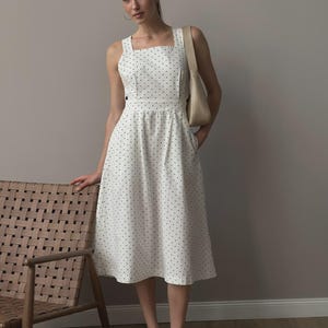 Puede incluir: Vestido midi blanco con estampado de lunares negros. El vestido presenta un escote cuadrado, tirantes anchos y un corpi&ntilde;o ajustado que fluye hacia una falda acampanada. Se lleva un bolso de hombro beige con el vestido. La persona lleva zapatos negros.