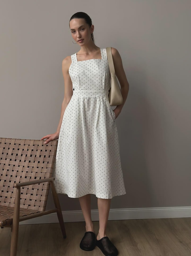 Puede incluir: Vestido midi blanco con estampado de lunares negros. El vestido presenta un escote cuadrado, tirantes anchos y un corpi&ntilde;o ajustado que fluye hacia una falda acampanada. Se lleva un bolso de hombro beige con el vestido. La persona lleva zapatos negros.