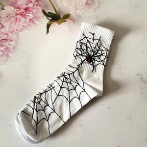Peut inclure: Chaussettes blanches avec une toile d'araignée noire et un motif d'araignée rouge.