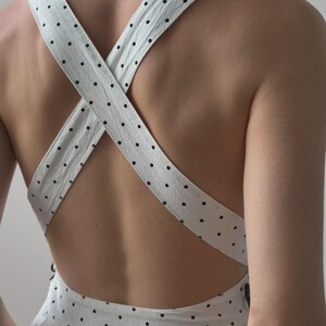 Puede incluir: Vestido blanco con lunares negros. El vestido presenta tirantes cruzados en la espalda. Las correas y la tela tienen una apariencia texturizada. Un vestido de estilo veraniego o primaveral.
