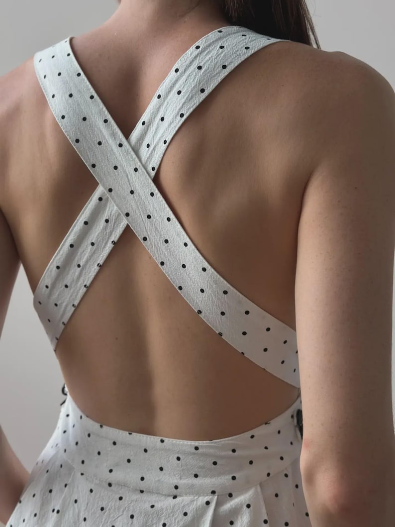 Puede incluir: Vestido blanco con lunares negros. El vestido presenta tirantes cruzados en la espalda. Las correas y la tela tienen una apariencia texturizada. Un vestido de estilo veraniego o primaveral.