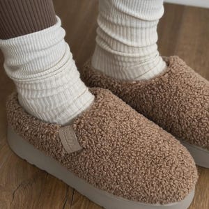 Könnte beinhalten: Braune, flauschige Hausschuhe mit einer hellbeigen Sohle. Die Hausschuhe haben ein weiches, strukturiertes Aussehen. Das Bild zeigt auch cremefarbene, gerippte Socken und braune Leggings.
